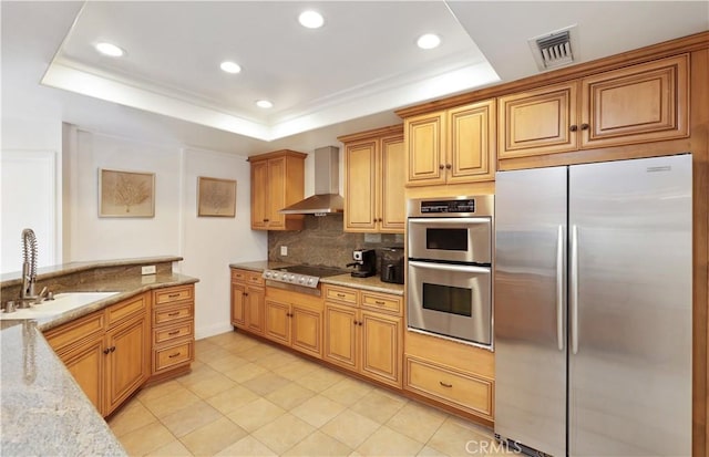 kitchen with stainless steel appliances, a sink, light stone countertops, wall chimney exhaust hood, and a raised ceiling