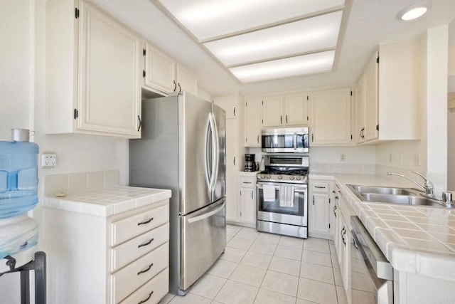 kitchen with tile countertops, stainless steel appliances, light tile patterned flooring, a sink, and white cabinetry