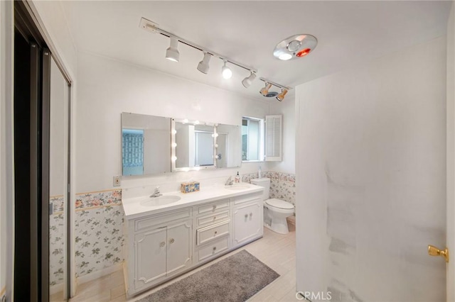 bathroom featuring toilet, double vanity, a sink, and wainscoting