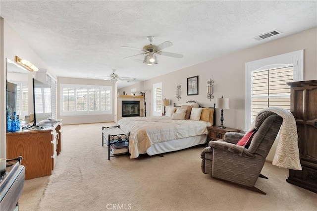 bedroom with light carpet, visible vents, a glass covered fireplace, ceiling fan, and a textured ceiling