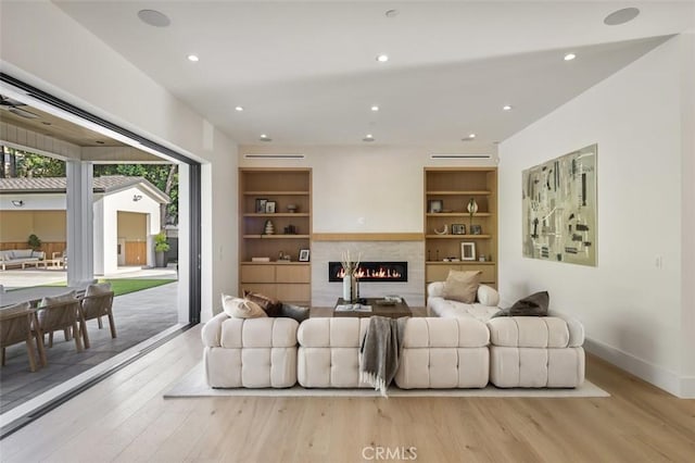 living area with built in features, recessed lighting, a warm lit fireplace, and light wood-style flooring