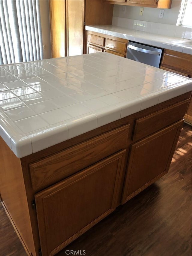 kitchen featuring brown cabinetry, tile countertops, dark wood-type flooring, a center island, and stainless steel dishwasher