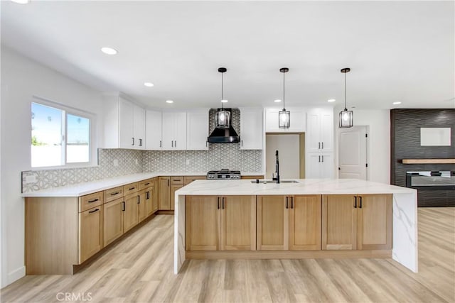 kitchen featuring wall chimney range hood, a sink, a center island with sink, and white cabinets