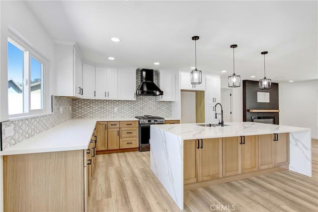 kitchen featuring stainless steel stove, a sink, white cabinets, wall chimney range hood, and an island with sink