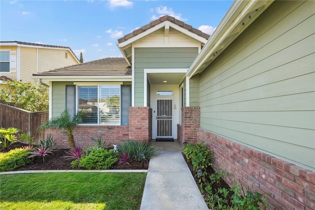 view of exterior entry with a tile roof, fence, a lawn, and brick siding