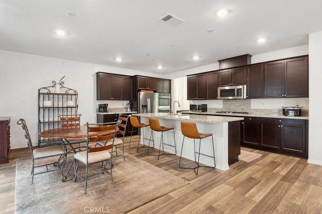 kitchen featuring a center island with sink, stainless steel appliances, visible vents, dark brown cabinetry, and light wood-type flooring