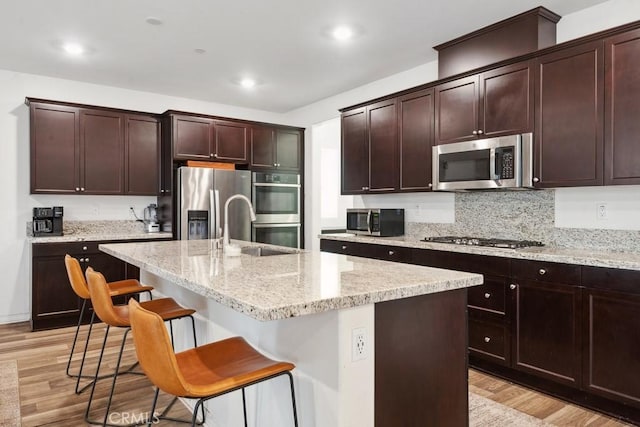 kitchen with a center island with sink, a breakfast bar area, appliances with stainless steel finishes, light stone countertops, and light wood-type flooring