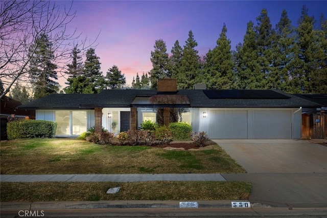 ranch-style house featuring a yard, a chimney, solar panels, an attached garage, and driveway