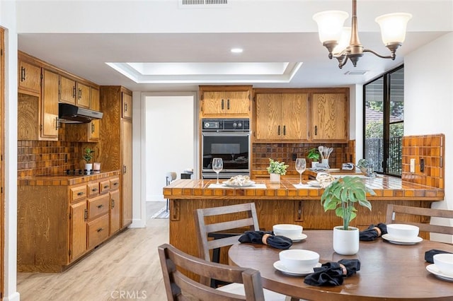 kitchen with under cabinet range hood, a peninsula, brown cabinets, a tray ceiling, and pendant lighting