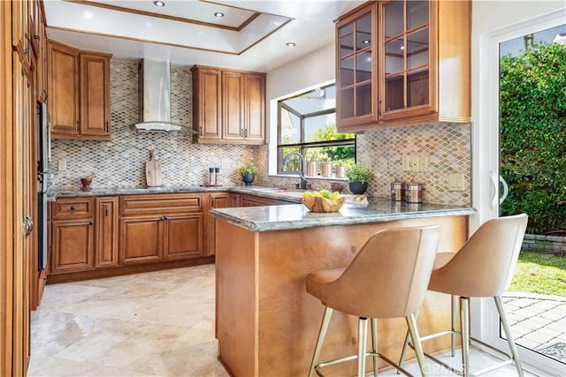 kitchen featuring glass insert cabinets, brown cabinets, wall chimney range hood, and a peninsula