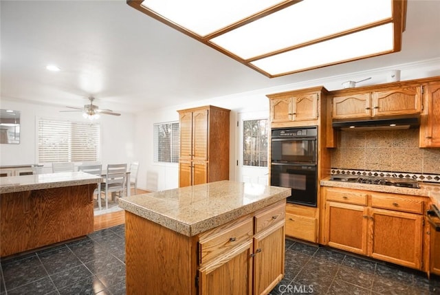 kitchen with under cabinet range hood, stainless steel gas cooktop, dobule oven black, a ceiling fan, and tasteful backsplash