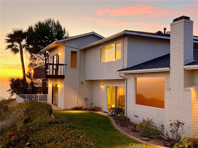 back of property at dusk featuring a balcony, a lawn, and stucco siding