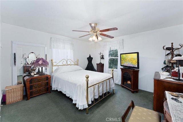 bedroom with a lit fireplace, dark carpet, and a ceiling fan