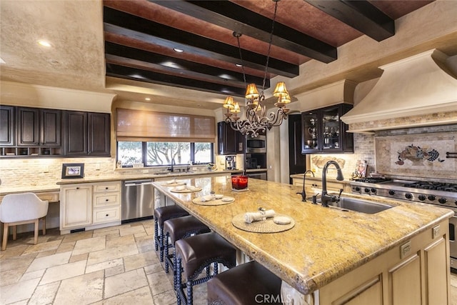 kitchen with appliances with stainless steel finishes, glass insert cabinets, a kitchen island with sink, a sink, and premium range hood