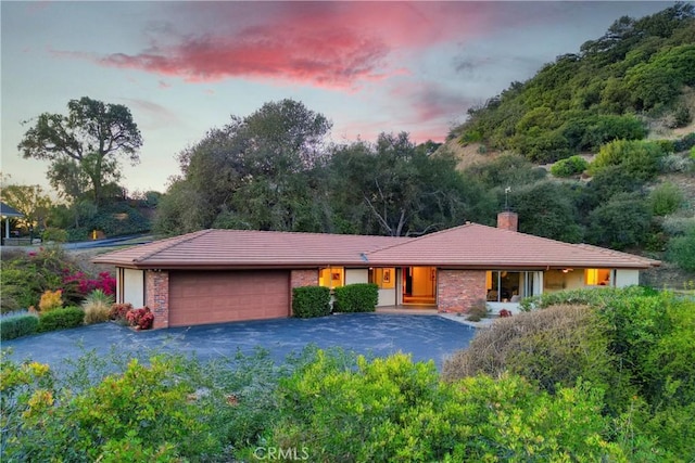 ranch-style house with aphalt driveway, an attached garage, a chimney, a tile roof, and brick siding