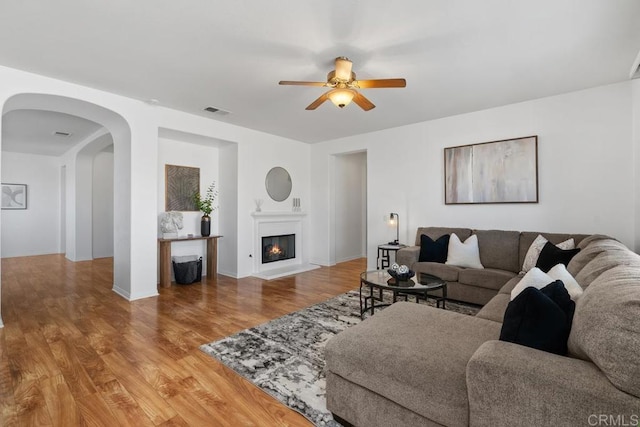 living area with arched walkways, visible vents, a ceiling fan, a glass covered fireplace, and wood finished floors