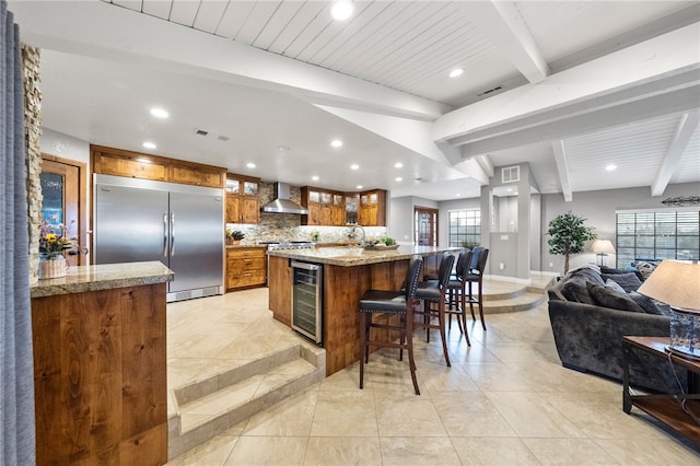 kitchen with wine cooler, beamed ceiling, stainless steel built in fridge, brown cabinets, and wall chimney exhaust hood