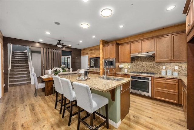 kitchen featuring light stone counters, under cabinet range hood, appliances with stainless steel finishes, light wood-type flooring, and a kitchen bar