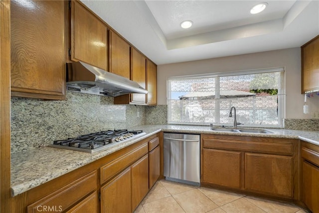 kitchen with under cabinet range hood, a sink, appliances with stainless steel finishes, brown cabinets, and a raised ceiling