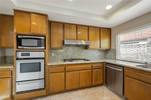 kitchen with under cabinet range hood, appliances with stainless steel finishes, brown cabinets, a warming drawer, and a raised ceiling