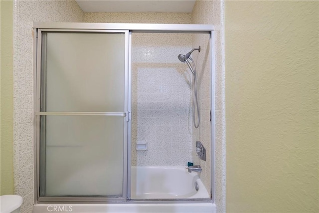 bathroom featuring a textured wall, shower / bath combination with glass door, and toilet