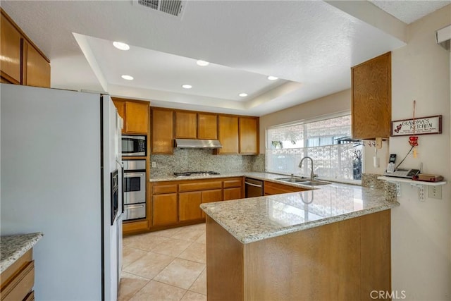 kitchen with a peninsula, a sink, appliances with stainless steel finishes, light stone countertops, and a raised ceiling