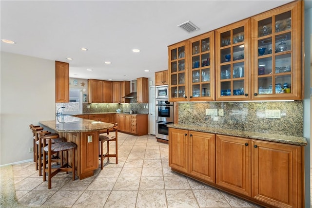 kitchen with wall chimney exhaust hood, glass insert cabinets, brown cabinetry, and light stone counters