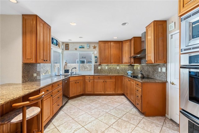 kitchen with brown cabinets, wall chimney exhaust hood, light stone counters, and stainless steel appliances