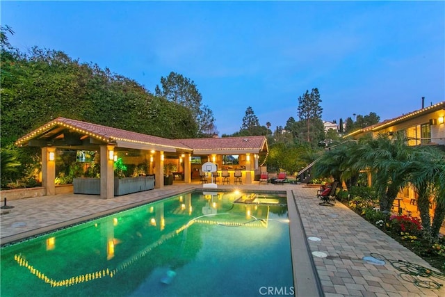 pool at dusk with area for grilling, a patio, an outdoor bar, and a pool with connected hot tub