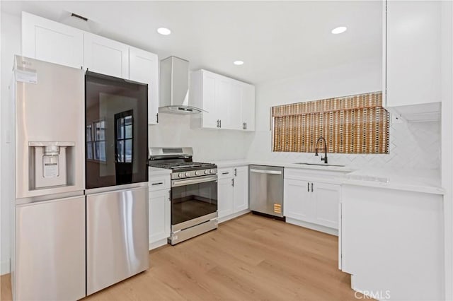kitchen with light wood finished floors, appliances with stainless steel finishes, white cabinets, wall chimney exhaust hood, and a sink