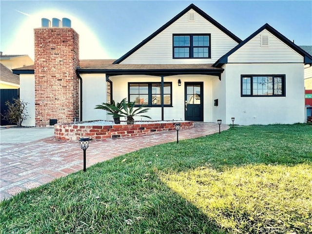 view of front of home with a front lawn, roof with shingles, covered porch, stucco siding, and a chimney