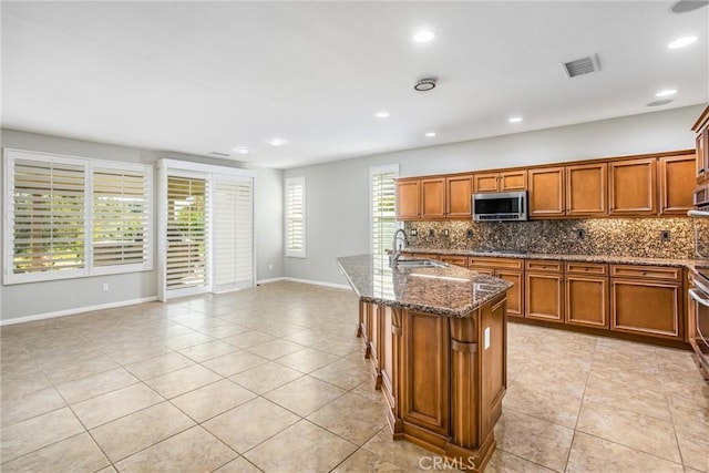 kitchen featuring brown cabinets, a kitchen island with sink, stainless steel microwave, and visible vents