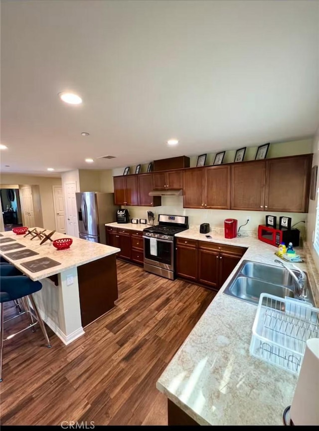 kitchen with under cabinet range hood, stainless steel appliances, dark wood-type flooring, a sink, and a kitchen breakfast bar