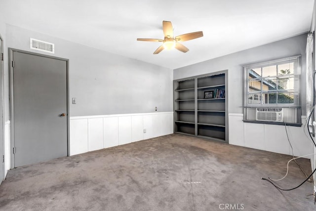 empty room featuring built in shelves, a wainscoted wall, carpet floors, visible vents, and a ceiling fan