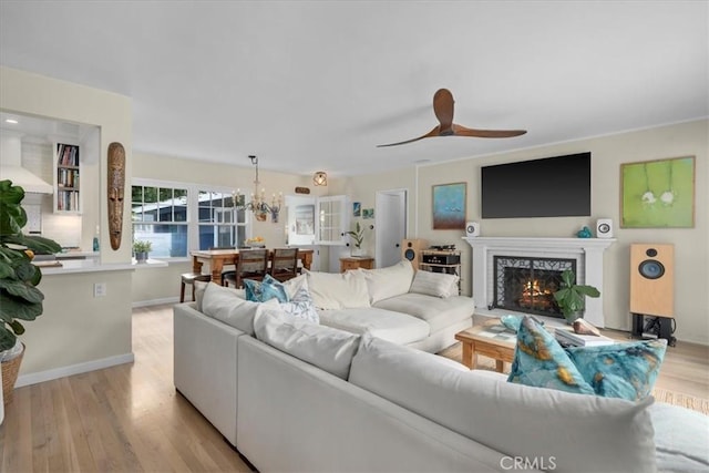 living room featuring baseboards, a lit fireplace, light wood finished floors, and ceiling fan with notable chandelier