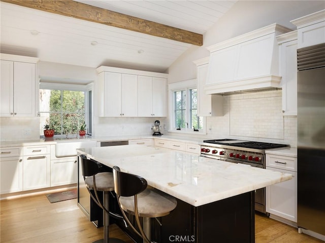 kitchen with light stone counters, custom range hood, lofted ceiling with beams, appliances with stainless steel finishes, and white cabinets