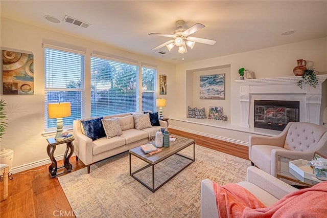 living room featuring ceiling fan, wood finished floors, visible vents, baseboards, and a glass covered fireplace