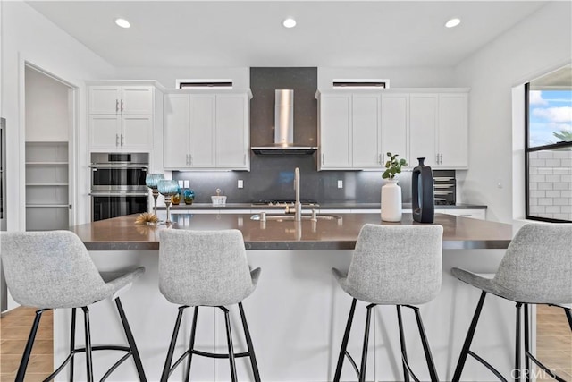 kitchen featuring stainless steel double oven, white cabinetry, wall chimney range hood, an island with sink, and dark countertops