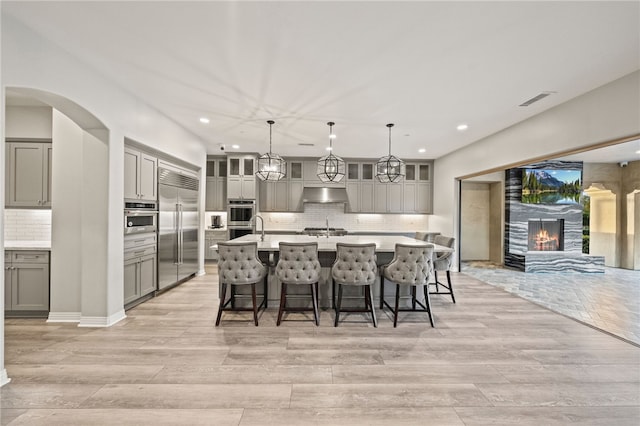 kitchen featuring an island with sink, appliances with stainless steel finishes, light countertops, under cabinet range hood, and pendant lighting