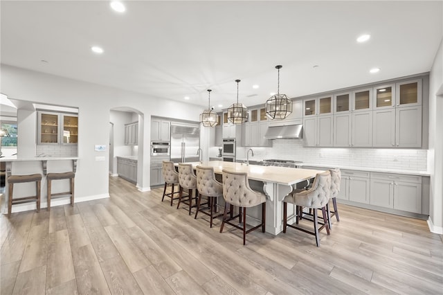 kitchen featuring light countertops, hanging light fixtures, glass insert cabinets, a large island with sink, and under cabinet range hood