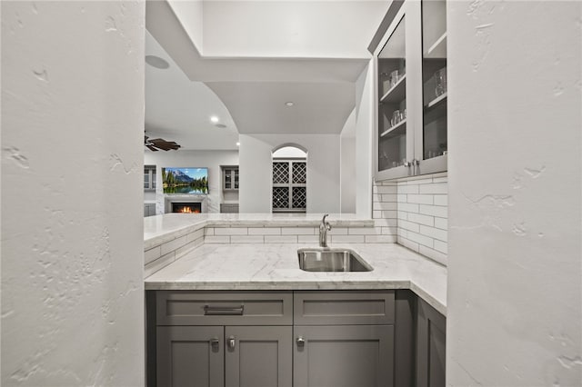 kitchen featuring light stone counters, gray cabinetry, a sink, decorative backsplash, and glass insert cabinets