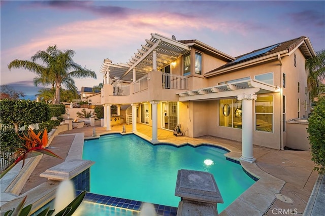 back of property at dusk with a fenced in pool, stucco siding, roof mounted solar panels, a balcony, and a pergola