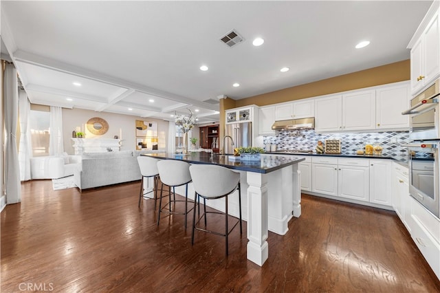 kitchen featuring white cabinets, an island with sink, dark countertops, a breakfast bar, and under cabinet range hood