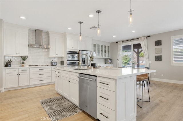 kitchen with stainless steel appliances, light countertops, a kitchen island with sink, white cabinetry, and wall chimney exhaust hood