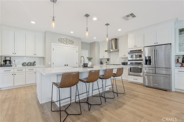 kitchen featuring wall chimney exhaust hood, appliances with stainless steel finishes, hanging light fixtures, a kitchen island with sink, and light countertops