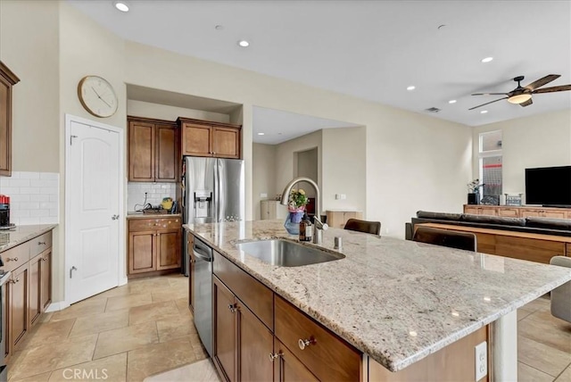 kitchen featuring light stone counters, brown cabinets, appliances with stainless steel finishes, a sink, and an island with sink