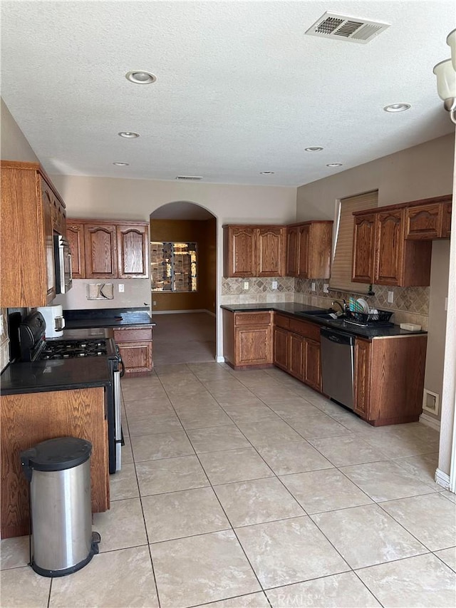 kitchen featuring arched walkways, stainless steel appliances, visible vents, brown cabinetry, and dark countertops