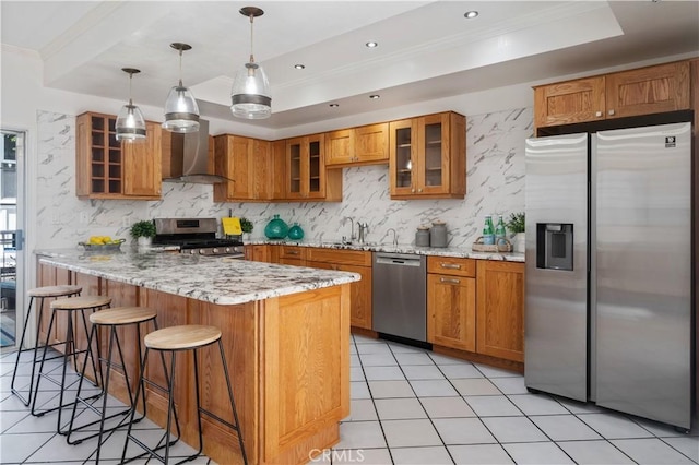 kitchen featuring decorative light fixtures, stainless steel appliances, a raised ceiling, glass insert cabinets, and wall chimney exhaust hood