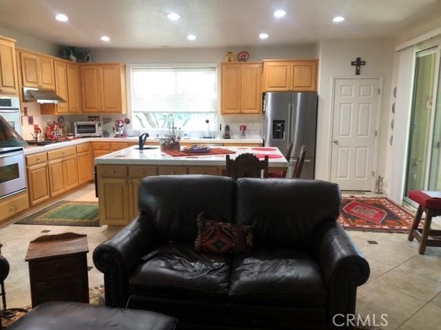 kitchen featuring appliances with stainless steel finishes, a kitchen island with sink, light countertops, light brown cabinetry, and under cabinet range hood