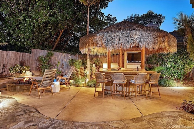 patio terrace at dusk with an outdoor fire pit, a fenced backyard, a grill, outdoor dry bar, and a gazebo